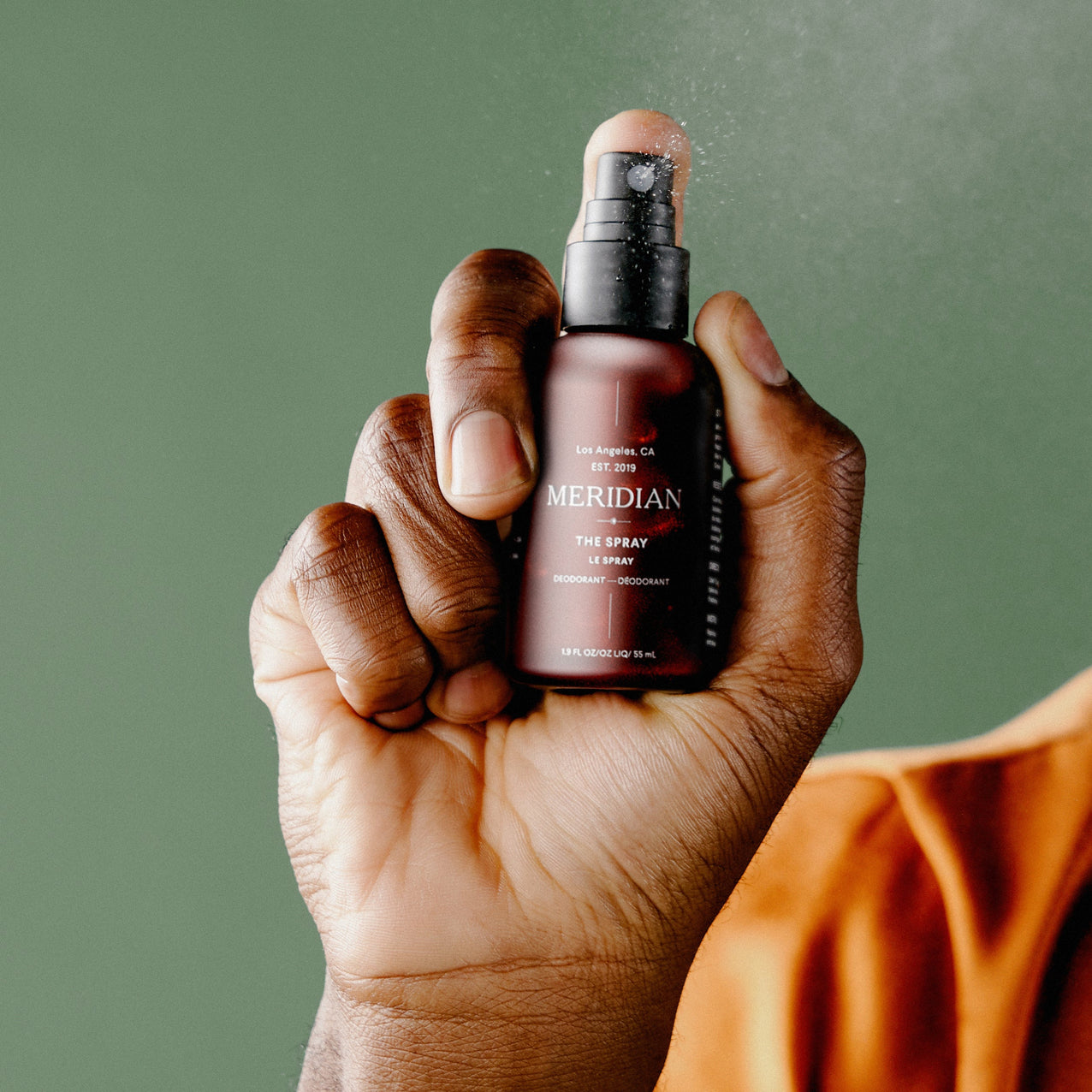 Close-up of a hand holding and spraying a Meridian branded deodorant spray bottle against a green background.