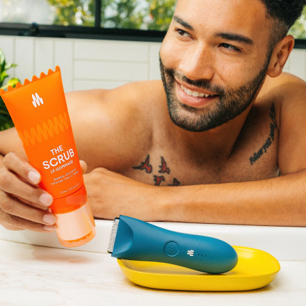 Man holding an orange tube of Meridian The Scrub exfoliating gel next to a blue electric trimmer placed on a yellow tray.