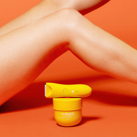 Close-up of smooth legs with a yellow electric grooming device and a container of The Pads grooming product on an orange background.