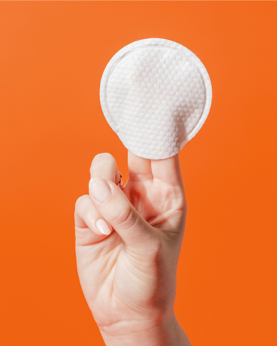Hand holding a white round cotton pad against an orange background.