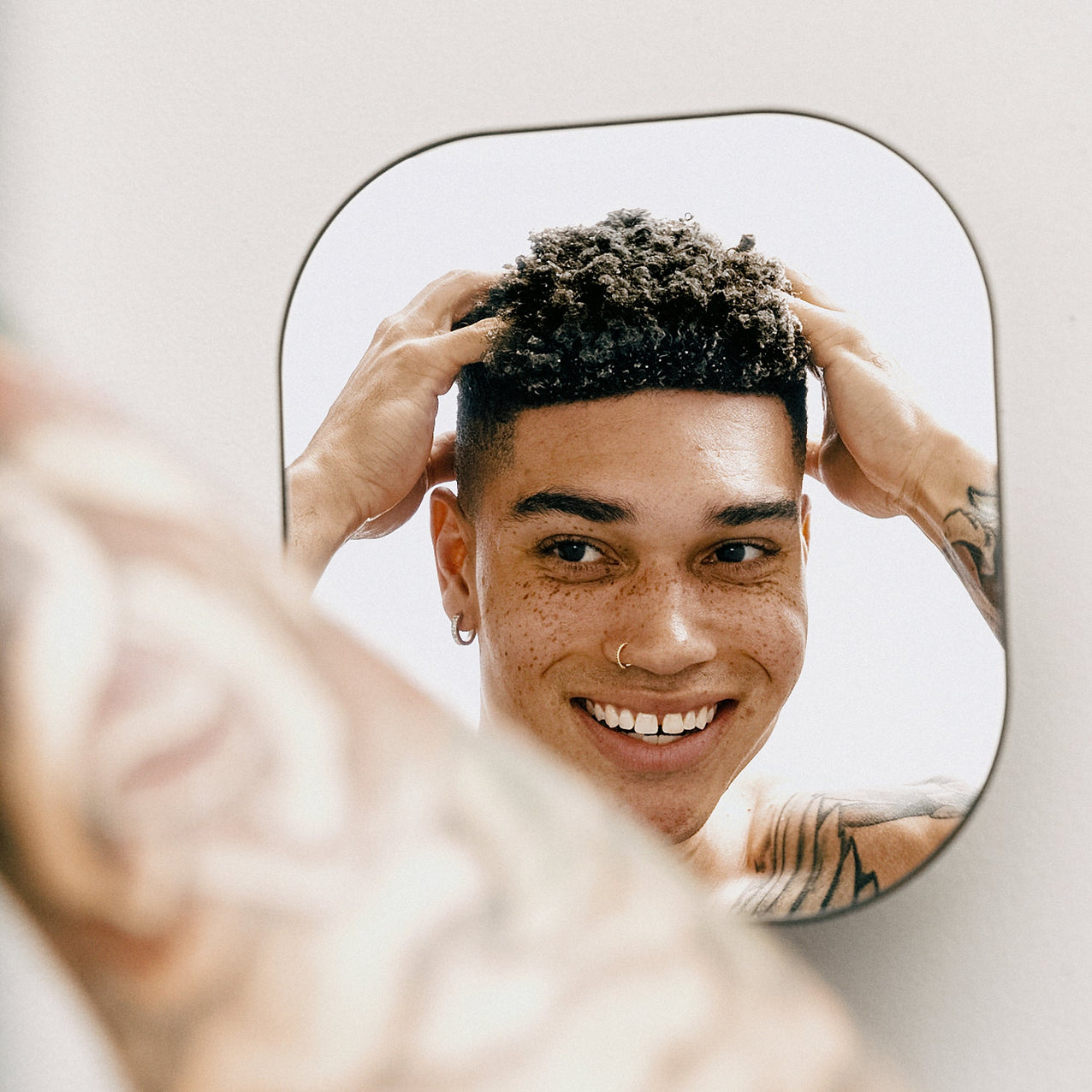 Smiling young man with curly hair and tattoos styling his hair while looking in the mirror.