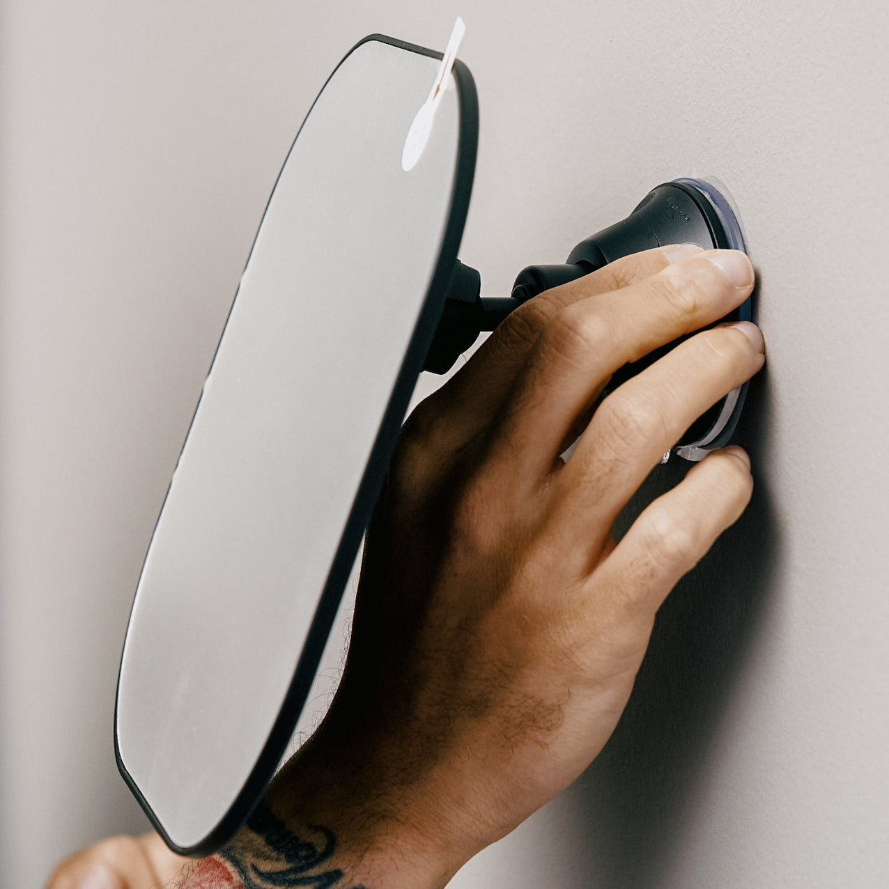 Hand with a tattoo adjusting a suction cup mirror mounted on a wall.