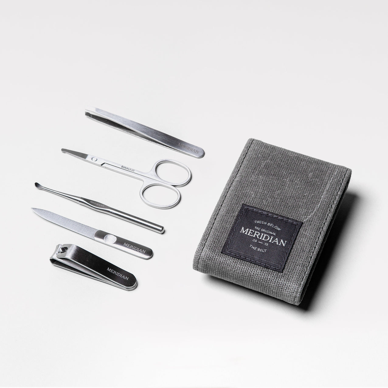 Men's grooming kit with stainless steel nail clippers, scissors, tweezers, nail file, and cuticle pusher next to a grey textured pouch labeled Meridian.