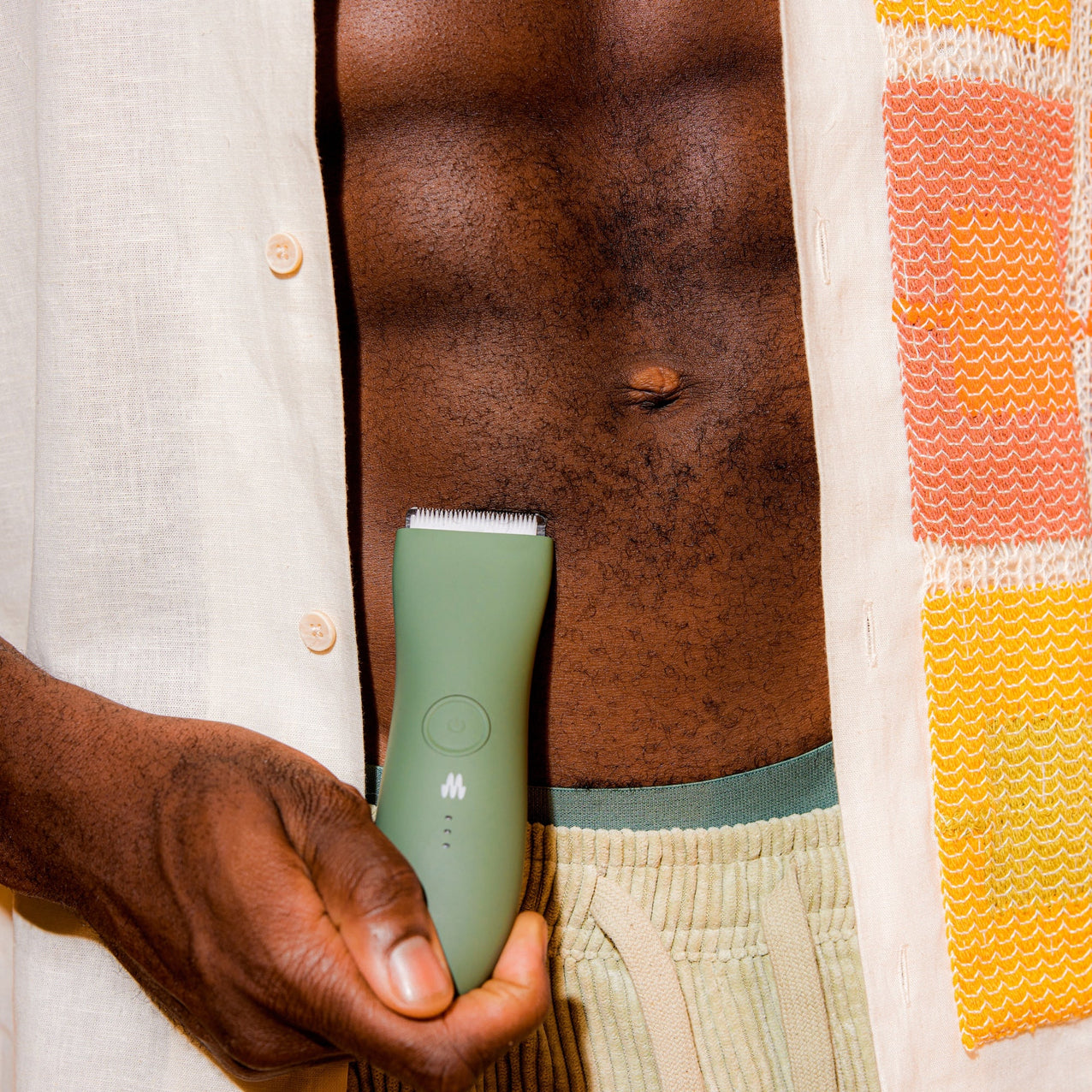 Handheld green facial cleansing brush on dark skin with open beige shirt and patterned orange towel, skincare routine tool.