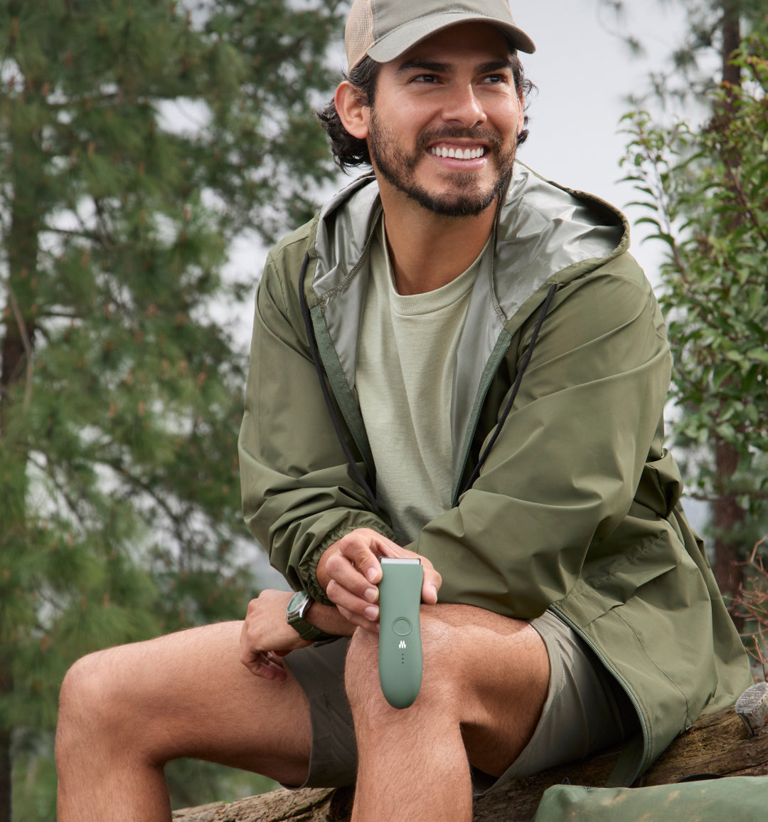 Man wearing olive green rain jacket, khaki shorts, and beige cap, sitting outdoors on log with wireless earbud case in hand